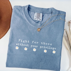 May include: A blue Comfort Colors t-shirt with the text "Fight for those without your privilege" and daisy graphics. A gold necklace with a pendant is on the shirt. A brown hat is in the upper left corner.