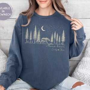 May include: A denim blue crewneck sweatshirt featuring a graphic of a bear and cub in a forest under a crescent moon and stars. The text reads Mama bear, Emily, and Oliver with a heart symbol. The design is printed in a light cream color.