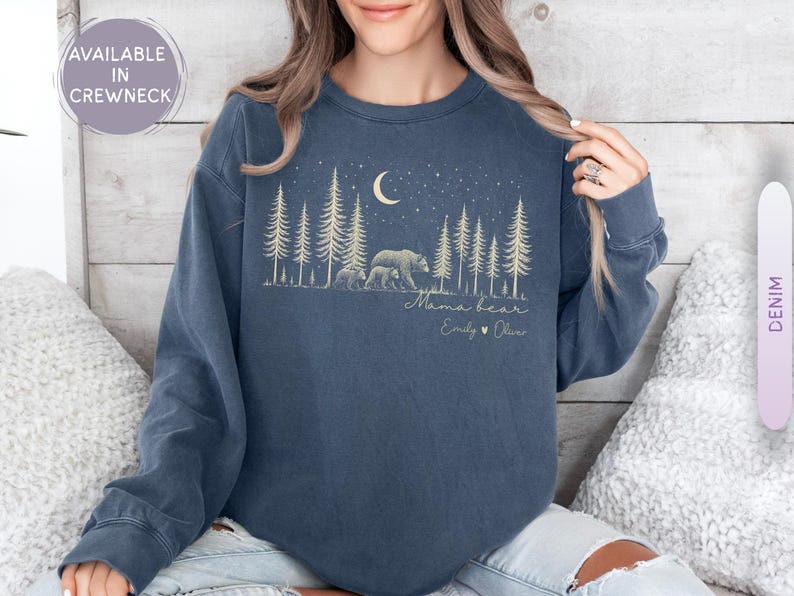 May include: A denim blue crewneck sweatshirt featuring a graphic of a bear and cub in a forest under a crescent moon and stars. The text reads Mama bear, Emily, and Oliver with a heart symbol. The design is printed in a light cream color.
