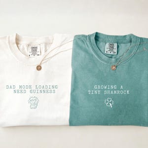 K&ouml;nnte beinhalten: Zwei T-Shirts, eines in Elfenbein und eines in Hellgr&uuml;n, mit Text und Grafiken. Das elfenbeinfarbene Shirt tr&auml;gt die Aufschrift "DAD MODE LOADING NEED GUINNESS" mit einer Bierkrug-Grafik. Das hellgr&uuml;ne Shirt tr&auml;gt die Aufschrift "GROWING A TINY SHAMROCK" mit einem Kleeblatt. Beide haben eine runde Anh&auml;nger-Halskette.