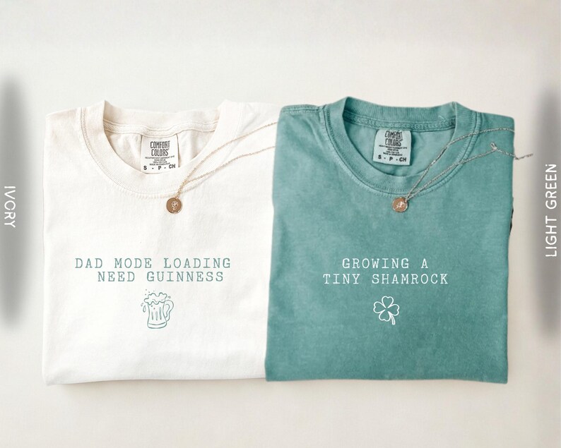K&ouml;nnte beinhalten: Zwei T-Shirts, eines in Elfenbein und eines in Hellgr&uuml;n, mit Text und Grafiken. Das elfenbeinfarbene Shirt tr&auml;gt die Aufschrift "DAD MODE LOADING NEED GUINNESS" mit einer Bierkrug-Grafik. Das hellgr&uuml;ne Shirt tr&auml;gt die Aufschrift "GROWING A TINY SHAMROCK" mit einem Kleeblatt. Beide haben eine runde Anh&auml;nger-Halskette.