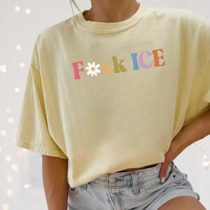 May include: A light yellow t-shirt with the text "F*ck ICE" in colorful letters. The "F" is a daisy, and the shirt is paired with denim shorts. The shirt is a banana color.