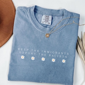 May include: A blue jean colored t-shirt with the text "KEEP THE IMMIGRANTS DEPORT THE RACISTS" in white, with daisy graphics. A gold necklace and a brown hat are also visible.