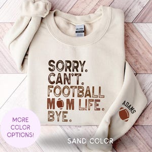 Peut inclure: Un sweat-shirt beige avec le texte "Sorry. Can't. Football Mom Life. Bye." en lettres brunes. Le texte est dans une police de caractères de style vintage. Un graphique de football brun est au-dessus du mot "Mom". Le texte "Adams" est sur la manche du sweat-shirt avec un graphique de football brun à côté.
