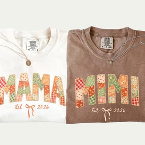 Peut inclure: Deux t-shirts, un ivoire et un marron, chacun avec un motif patchwork épelant "MAMA" et "MIMI". Les chemises ont un nœud et le texte "Est 2026" en dessous. Chaque chemise a un collier.