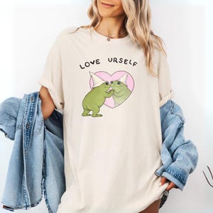 Pu&ograve; includere: T-shirt color avorio con la scritta "LOVE URSELF" sopra una grafica di due rane verdi che si baciano all'interno di un cuore rosa. Le rane indossano cappelli bianchi. Una giacca di jeans blu &egrave; drappeggiata sulle braccia.