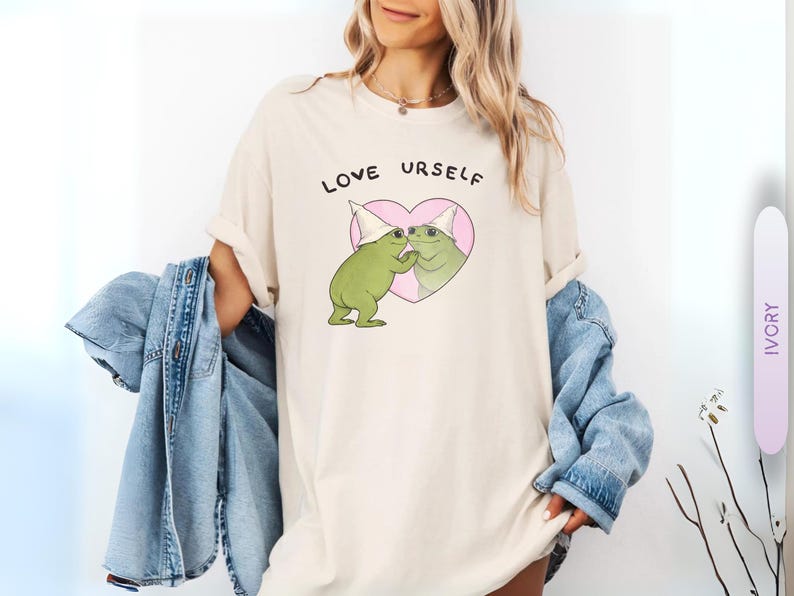 Pu&ograve; includere: T-shirt color avorio con la scritta "LOVE URSELF" sopra una grafica di due rane verdi che si baciano all'interno di un cuore rosa. Le rane indossano cappelli bianchi. Una giacca di jeans blu &egrave; drappeggiata sulle braccia.