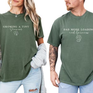 May include: Two olive green t-shirts. One says "Growing a Tiny Shamrock" with a shamrock graphic. The other says "Dad Mode Loading. need Guinness" with a beer graphic. Both shirts have short sleeves and a relaxed fit.