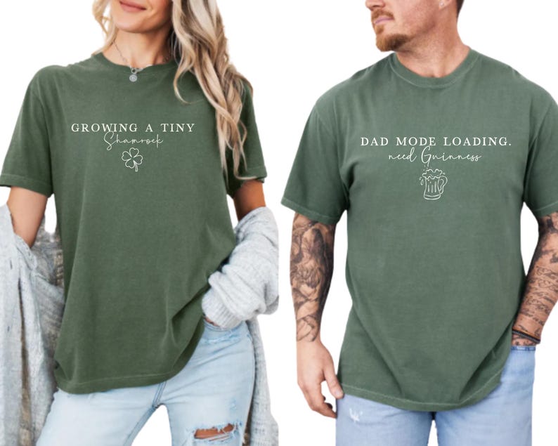 May include: Two olive green t-shirts. One says "Growing a Tiny Shamrock" with a shamrock graphic. The other says "Dad Mode Loading. need Guinness" with a beer graphic. Both shirts have short sleeves and a relaxed fit.