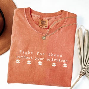 May include: Terracotta-colored t-shirt with the text "Fight for those without your privilege" and daisy graphics. A gold necklace and a brown hat are also visible. The shirt is a Comfort Colors brand.