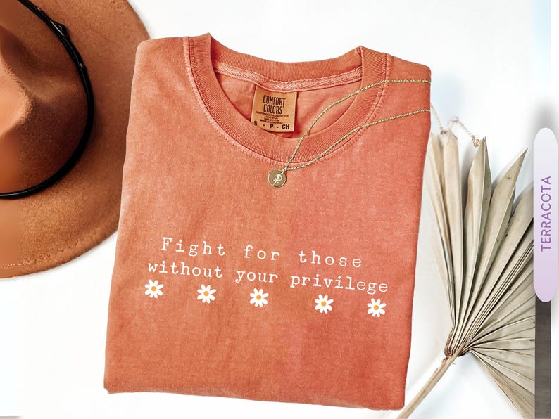 May include: Terracotta-colored t-shirt with the text "Fight for those without your privilege" and daisy graphics. A gold necklace and a brown hat are also visible. The shirt is a Comfort Colors brand.