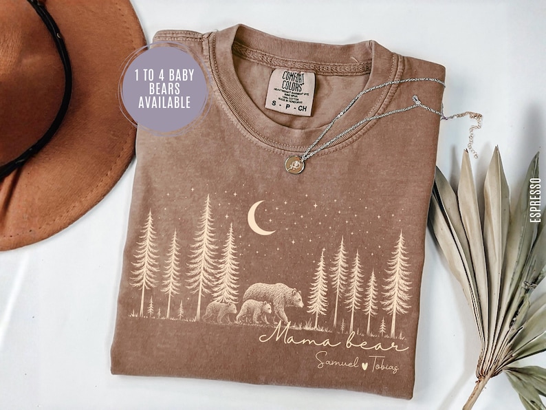 May include: An espresso-colored t-shirt featuring a graphic of a forest with pine trees, a crescent moon, stars, and a mother bear with two cubs. The text reads "Mama bear" and "Samuel Tobias" with a heart. A circular label says "1 TO 4 BABY BEARS AVAILABLE".