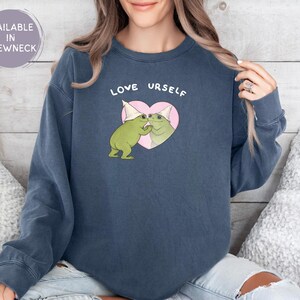 Pu&ograve; includere: Una felpa girocollo blu navy con la scritta "LOVE URSELF" sopra una grafica di due rane verdi con cappelli da festa che si abbracciano all'interno di un cuore rosa. La felpa &egrave; disponibile con girocollo.