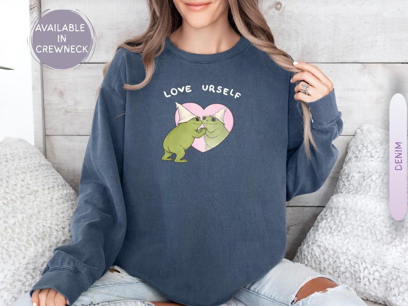 Pu&ograve; includere: Una felpa girocollo blu navy con la scritta "LOVE URSELF" sopra una grafica di due rane verdi con cappelli da festa che si abbracciano all'interno di un cuore rosa. La felpa &egrave; disponibile con girocollo.