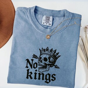 Peut inclure: Un t-shirt bleu clair Comfort Colors à col rond, orné d'un motif noir d'un crâne couronné avec une flèche traversant son orbite. Le texte "No kings" est en dessous. Un collier doré est posé sur le t-shirt.