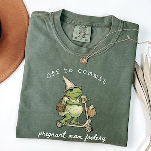 Peut inclure: T-shirt vert avec l'inscription "Off to commit pregnant mom foolery" et une grenouille de dessin animé sur un scooter. La grenouille porte un chapeau et tient une tasse de café. Un chapeau marron et un collier sont également visibles.