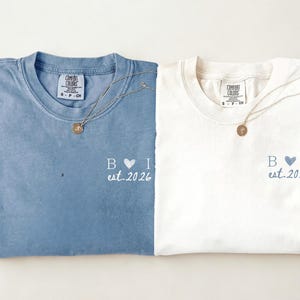 Peut inclure: Deux t-shirts pliés, un bleu et un ivoire, avec des colliers assortis. Chaque chemise porte les initiales "B I" avec un cœur et le texte "est.2026". Le t-shirt bleu est étiqueté "Bleu Jean" et le t-shirt ivoire est étiqueté "Ivory".