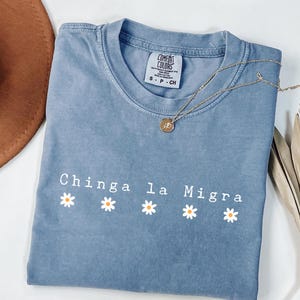 Peut inclure: Un t-shirt bleu jean avec l'inscription "Chinga la Migra" en lettres blanches et quatre motifs de marguerites. Un collier doré et un chapeau marron sont également présents.