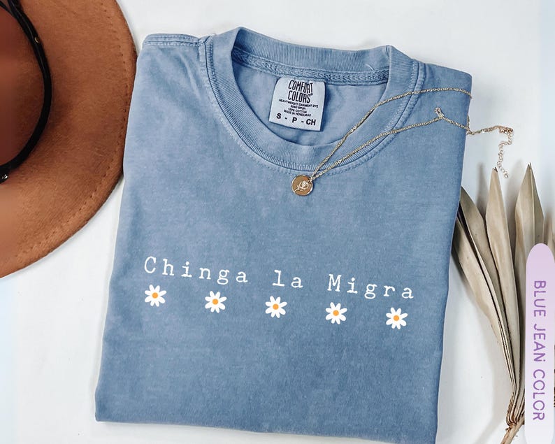 May include: A blue jean colored t-shirt with the phrase "Chinga la Migra" in white letters and four daisy graphics. A gold necklace and a brown hat are also in the image.