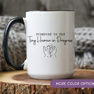 Peut inclure: Mug en céramique blanche avec une anse et un bord noirs. Le mug porte l'inscription "Promu Papa Tiny Human in Progress" en écriture noire, ainsi qu'un dessin au trait de mains berçant les pieds d'un bébé. Le mug est posé sur une surface en bois.