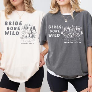 Puede incluir: Camisetas marfil y grises con el texto "BRIDE GONE WILD" y "GIRLS GONE WILD". Cada camiseta tiene un gr&aacute;fico de una escena de acampada y el texto "Emily's Bachelorette 2025 Lake Tahoe, CA".