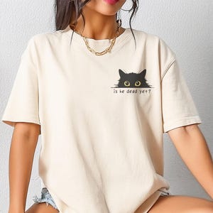 Peut inclure: T-shirt ivoire avec un motif de chat noir et le texte "is he dead yet ?". Le t-shirt a des manches courtes et une coupe décontractée, idéal pour un usage quotidien.