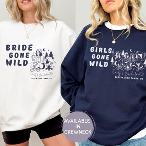 Puede incluir: Sudaderas blancas y azul marino con las frases "Bride Gone Wild" y "Girls Gone Wild" en azul marino. Cada sudadera tiene un gr&aacute;fico con tem&aacute;tica de camping y el texto "Emily's Bachelorette 2025 Lake Tahoe, CA."
