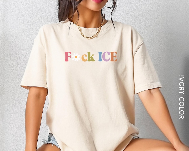 May include: Ivory-colored t-shirt with the text "F*ck ICE" in colorful, retro-style lettering. The shirt features a daisy graphic between the "F" and "ck". The shirt is a casual, short-sleeved design.