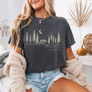 May include: A charcoal gray short-sleeve t-shirt featuring a graphic print of a forest with pine trees, a crescent moon, and stars. The design includes a bear with two cubs, the text "Mama bear," and the names "Emily" and "Oliver" with a heart.