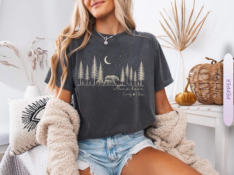 May include: A charcoal gray short-sleeve t-shirt featuring a graphic print of a forest with pine trees, a crescent moon, and stars. The design includes a bear with two cubs, the text "Mama bear," and the names "Emily" and "Oliver" with a heart.