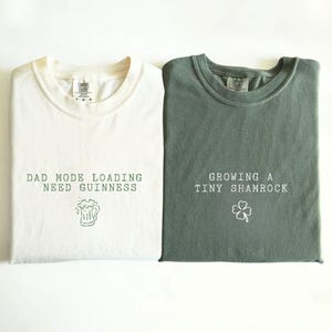 K&ouml;nnte beinhalten: Zwei gefaltete T-Shirts, eines in Elfenbein und eines in Moosgr&uuml;n. Das elfenbeinfarbene Shirt hat den Text "DAD MODE LOADING NEED GUINNESS" mit einer Bierkrug-Grafik. Das moosgr&uuml;ne Shirt sagt "GROWING A TINY SHAMROCK" mit einem Kleeblatt.