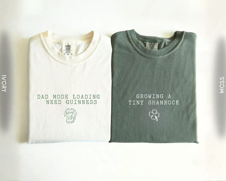 K&ouml;nnte beinhalten: Zwei gefaltete T-Shirts, eines in Elfenbein und eines in Moosgr&uuml;n. Das elfenbeinfarbene Shirt hat den Text "DAD MODE LOADING NEED GUINNESS" mit einer Bierkrug-Grafik. Das moosgr&uuml;ne Shirt sagt "GROWING A TINY SHAMROCK" mit einem Kleeblatt.