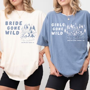 Puede incluir: Dos camisetas holgadas, una marfil y otra azul vaquero, con los textos "BRIDE GONE WILD" y "GIRLS GONE WILD" en azul. Cada camiseta presenta un dise&ntilde;o de acampada y el texto "Emily's Bachelorette 2025 Lake Tahoe, CA."