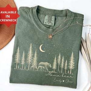 May include: A moss green crewneck t-shirt featuring a cream-colored graphic of a forest with pine trees, a crescent moon, stars, and a mother bear with two cubs. The text reads Mama bear, Emily, and Oliver. The shirt has a Comfort Colors label.