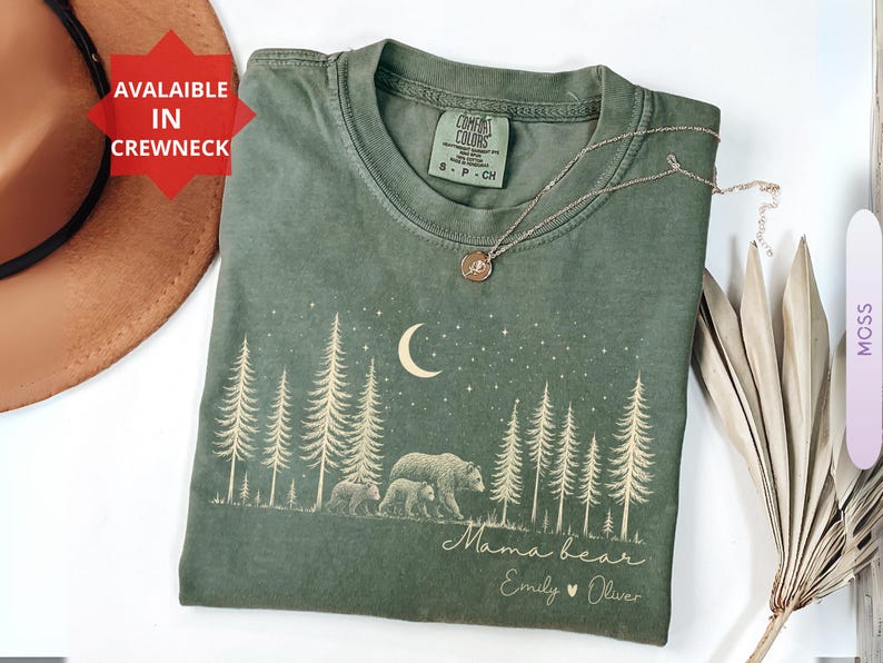 May include: A moss green crewneck t-shirt featuring a cream-colored graphic of a forest with pine trees, a crescent moon, stars, and a mother bear with two cubs. The text reads Mama bear, Emily, and Oliver. The shirt has a Comfort Colors label.