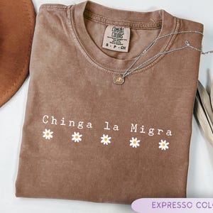 May include: A brown, short-sleeved t-shirt with the text "Chinga la Migra" in white, with daisy graphics. A silver necklace with a pendant rests on the shirt. The shirt is labeled "Expresso Color".