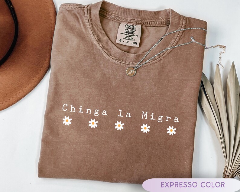May include: A brown, short-sleeved t-shirt with the text "Chinga la Migra" in white, with daisy graphics. A silver necklace with a pendant rests on the shirt. The shirt is labeled "Expresso Color".