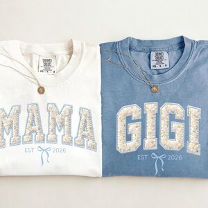 May include: Two crew neck t-shirts, one ivory and one blue, with the words "MAMA" and "GIGI" in a floral design. Each shirt has a matching necklace and the text "EST 2026".