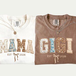 Peut inclure: Deux t-shirts pliés, un ivoire et un marron, chacun avec un motif floral pour le nom. Le t-shirt ivoire dit "MAMA" et le t-shirt marron dit "GIGI", tous deux avec "EST 2026" et un motif de nœud. Chaque chemise a un collier.