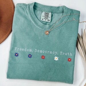 May include: A light green t-shirt with the words "Freedom, Democracy, Truth" and flower graphics. The shirt has a crew neck and short sleeves. A gold necklace with a pendant is draped on the shirt.