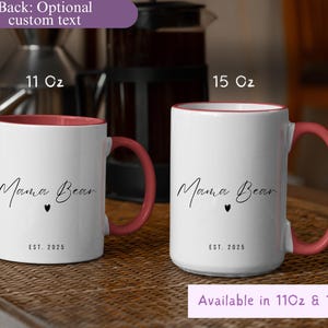 May include: Two white ceramic mugs with a pink interior and handle. The mugs have the text "Mama Bear" in a script font, a small heart, and "EST. 2025". One mug is 11 oz, the other is 15 oz. The mugs are on a woven surface.