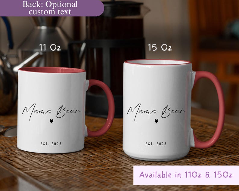 May include: Two white ceramic mugs with a pink interior and handle. The mugs have the text "Mama Bear" in a script font, a small heart, and "EST. 2025". One mug is 11 oz, the other is 15 oz. The mugs are on a woven surface.