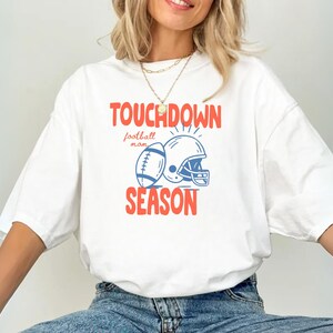 Puede incluir: Camiseta blanca con un gr&aacute;fico tem&aacute;tico de f&uacute;tbol. El dise&ntilde;o presenta un bal&oacute;n de f&uacute;tbol, un casco y las palabras "TOUCHDOWN SEASON" en rojo y azul. Tambi&eacute;n incluye la frase "football mom".