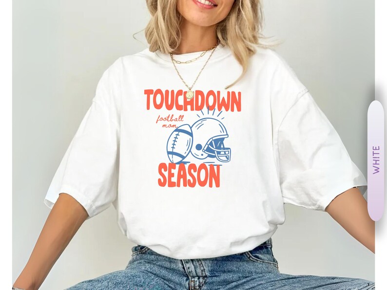 Puede incluir: Camiseta blanca con un gr&aacute;fico tem&aacute;tico de f&uacute;tbol. El dise&ntilde;o presenta un bal&oacute;n de f&uacute;tbol, un casco y las palabras "TOUCHDOWN SEASON" en rojo y azul. Tambi&eacute;n incluye la frase "football mom".