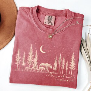 May include: A crimson short-sleeve t-shirt featuring a cream-colored graphic of a forest, a crescent moon, stars, and a mother bear with three cubs. The text reads Mama bear followed by the names River, Jade, and Bella with heart icons.