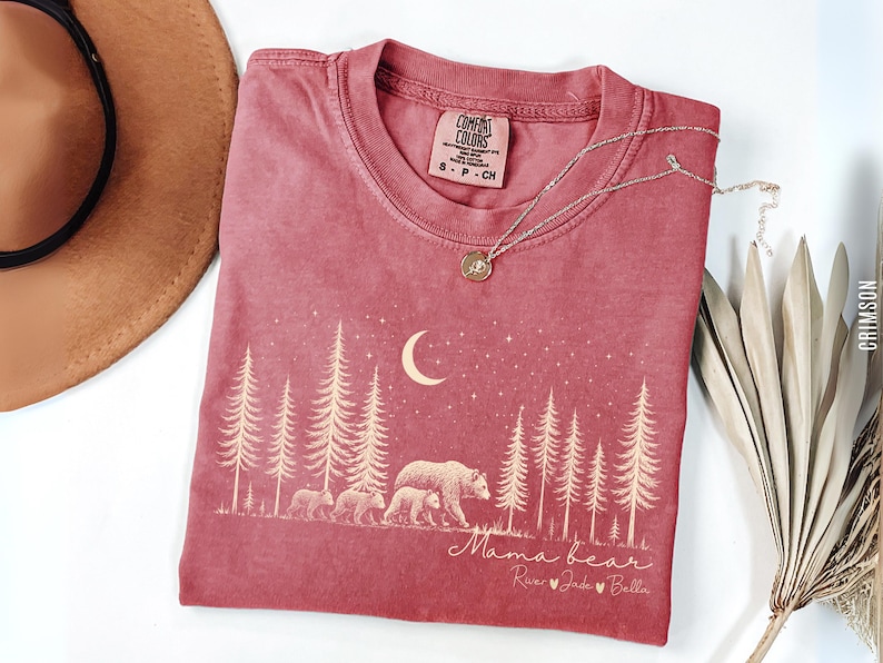 May include: A crimson short-sleeve t-shirt featuring a cream-colored graphic of a forest, a crescent moon, stars, and a mother bear with three cubs. The text reads Mama bear followed by the names River, Jade, and Bella with heart icons.