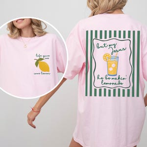 May include: A light pink t-shirt with a lemon graphic on the front and a lemonade graphic on the back. The front of the shirt says "life gave me some lemons." The back says "but my Jesus he be makin' lemonade."