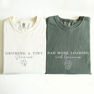 May include: Two folded t-shirts, one ivory and one moss green, are displayed. The ivory shirt has the text "GROWING A TINY Shamrock" with a shamrock graphic. The moss green shirt reads "DAD MODE LOADING. need Guinness" with a beer mug graphic.