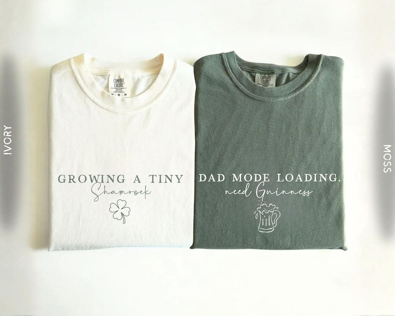 May include: Two folded t-shirts, one ivory and one moss green, are displayed. The ivory shirt has the text "GROWING A TINY Shamrock" with a shamrock graphic. The moss green shirt reads "DAD MODE LOADING. need Guinness" with a beer mug graphic.