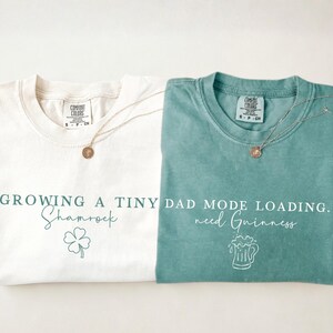 May include: Two t-shirts, one ivory and one light green, with text and graphics. The ivory shirt says "Growing a Tiny Shamrock" with a shamrock graphic. The light green shirt says "Dad Mode Loading. need Guinness" with a beer mug graphic.